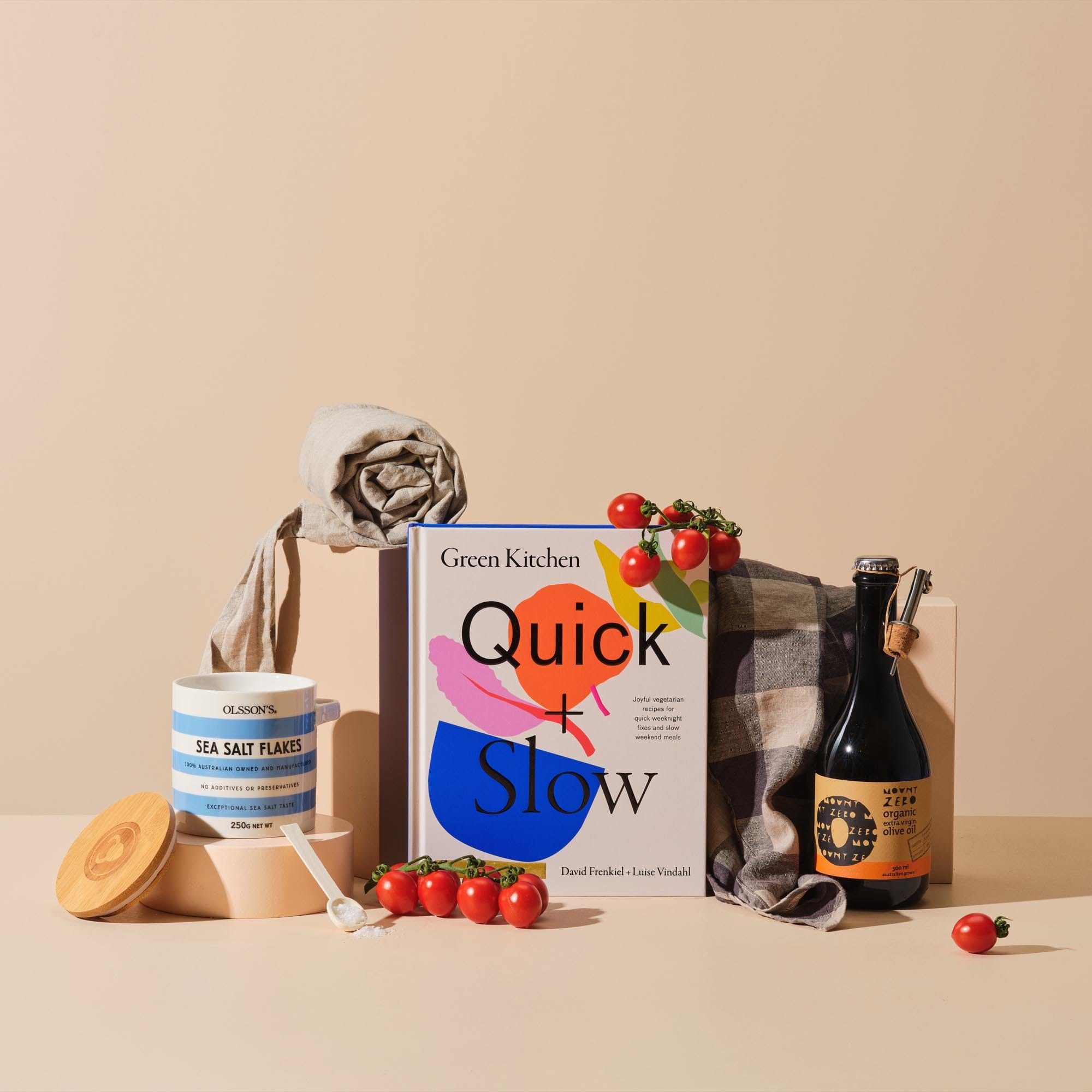This image displays the gift bundle 'Veg Out' featuring the cover of 'Quick and Slow', Mount Zero Olive Oil, Olsson's Sea Salt Flakes, Linen Tea Towel and Apron, accessorised with red cherry tomatoes. 