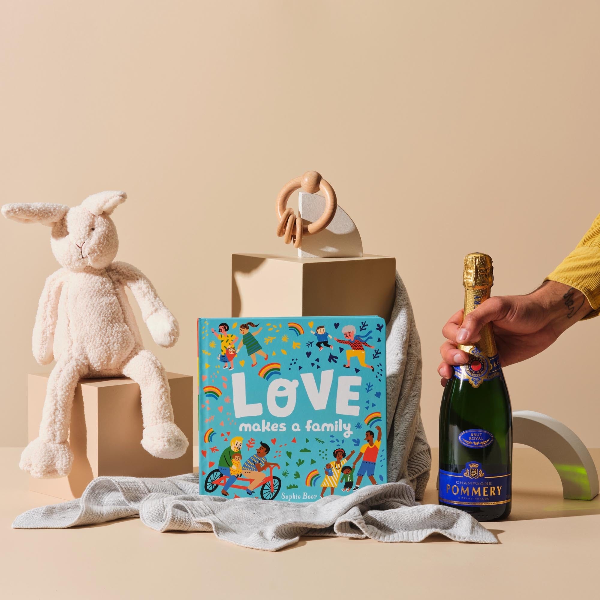 This is the Champagne Baby Showers gift bundle from Handsel. The image displays Sophie Beer's Love Makes a Family, Purebaby Essentials Blanket and Teething Ring, Bonnie the Bunny Toy and Champagne Pommery Brut Royal